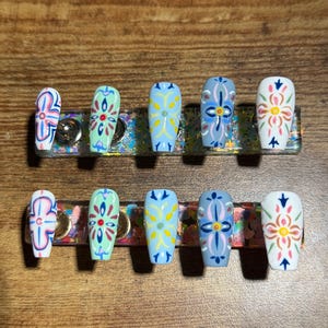 May include: A set of ten coffin-shaped false nails. Each nail features a different colorful floral design on a white, light blue, or mint green base. The designs include pink, yellow, and blue accents. The nails are displayed on a clear holder.