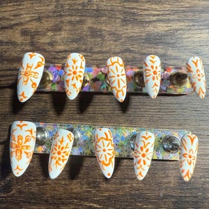 May include: A set of white, almond-shaped false nails with orange floral designs. The nails are arranged in two rows on a decorative holder with iridescent glitter. The background is a dark wood surface.