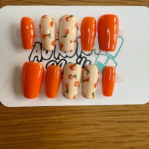 May include: A set of press-on nails in a variety of shapes. Some nails are solid orange, while others are a light peach color with a pattern of small orange cherries and green leaves. The nails are arranged on a white card.