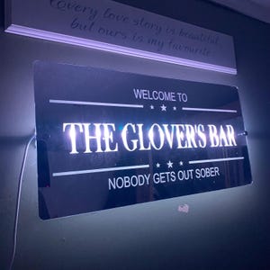 Peut inclure: Une enseigne noire et blanche avec des lumières blanches qui indique "Bienvenue au Glover's Bar" et "Personne ne sort sobre".
