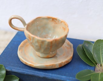 Könnte beinhalten: Ein hellgrünes und orangefarbenes Keramik-Teetassen- und Untertassen-Set mit einer strukturierten, unebenen Oberfläche. Das Set steht auf einem blauen Buch mit dem Text "Living English Structure" auf dem Rücken.