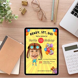 K&ouml;nnte beinhalten: Eine digitale Einladung auf einem Tablet zum 6. Geburtstag von David, mit einem Cartoon-Flieger und Luftballons. Der gelbe Hintergrund tr&auml;gt den Text "READY, SET, GO!" und Veranstaltungsdetails. Weitere Gegenst&auml;nde auf der Holzoberfl&auml;che sind ein Laptop, ein Taschenrechner und Schreibwaren.