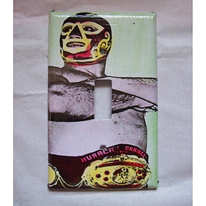 May include: A light switch cover featuring a wrestler wearing a colorful mask and a championship belt with the text "HURRACA RAMIREZ".