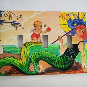 May include: A light switch plate featuring a vintage illustration of a mermaid with a green tail, holding an umbrella, and a cupid with a red heart. The background is a light pink and blue.