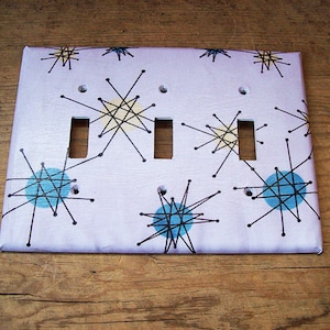 May include: A light purple light switch cover with a mid-century modern design featuring black and blue stars on a white background.