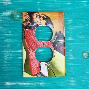 May include: A vintage-style light switch cover featuring a chimpanzee wearing a red and white checkered shirt and white shoes. The chimpanzee is sitting on a chair and holding a phone.