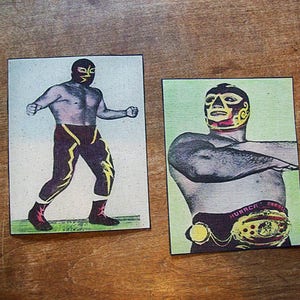 May include: Two vintage wrestling trading cards featuring masked wrestlers. The first card shows a wrestler in a black and yellow costume with a black mask. The second card shows a wrestler in a red and yellow costume with a black mask. Both cards have a green background.