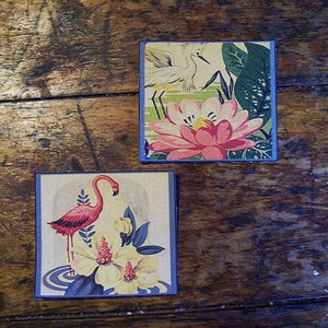 Può includere: Due cartoline vintage con illustrazioni di fiori e uccelli. La prima cartolina presenta un giglio d'acqua rosa con foglie verdi e un uccello bianco con un lungo becco. La seconda cartolina presenta un fenicottero rosa in piedi in uno stagno con un fiore bianco.