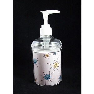 May include: A clear plastic soap dispenser with a white pump. The dispenser has a pink and white label with a mid-century modern design of blue and yellow stars.