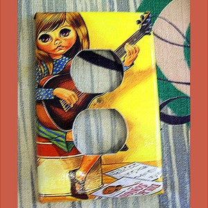 May include: A yellow and brown outlet cover with a picture of a girl with large eyes playing a guitar. The girl is wearing a blue and white polka dot shirt and a red, white, and blue skirt. The text "Songs of Joy" is visible on the sheet music in the bottom right corner.