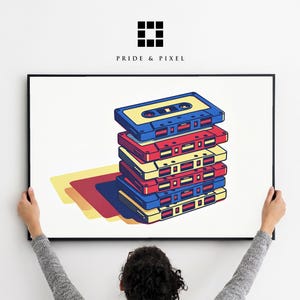 Puede incluir: Una impresión enmarcada con una pila de cintas de cassette retro en azul, rojo y amarillo. La obra de arte incluye el texto "PRIDE & PIXEL" en la parte superior. La imagen es sostenida por una persona.