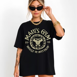 May include: Black t-shirt with a circular graphic that reads "MAUI'S GYM" in a beige color. The graphic features a cartoon character flexing, with the text "MUSCLE UP, BUTTERCUP" below. The shirt is worn by a person wearing sunglasses and gold jewelry.