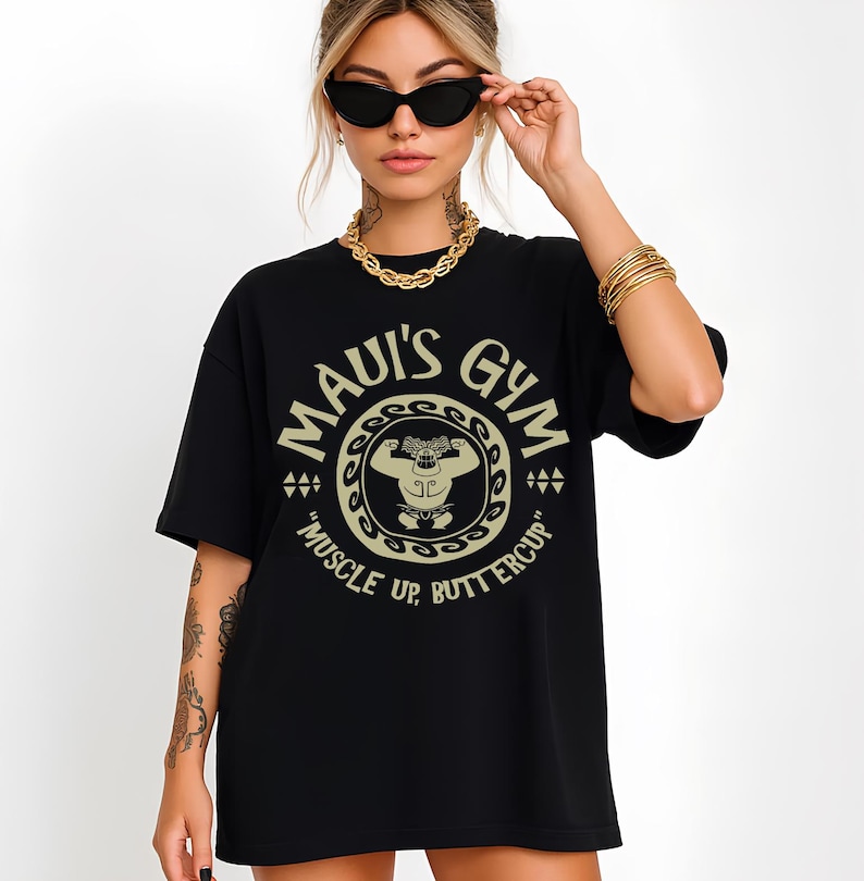 May include: Black t-shirt with a circular graphic that reads "MAUI'S GYM" in a beige color. The graphic features a cartoon character flexing, with the text "MUSCLE UP, BUTTERCUP" below. The shirt is worn by a person wearing sunglasses and gold jewelry.
