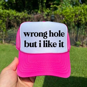 May include: A hot pink trucker hat with a white front panel. The text "wrong hole but i like it" is printed in black on the white panel. The hat has a mesh back and a curved brim.