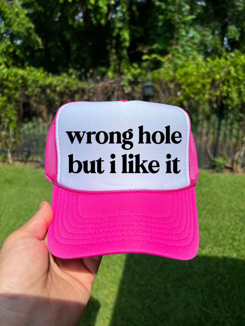 May include: A hot pink trucker hat with a white front panel. The text "wrong hole but i like it" is printed in black on the white panel. The hat has a mesh back and a curved brim.
