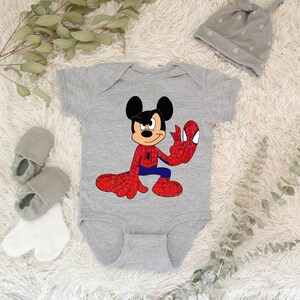 Op de afbeelding: Een grijze babybody met een cartoon Mickey Mouse-personage in een Spider-Man-kostuum. De body heeft korte mouwen en een drukknoopsluiting. Een grijze beanie, grijze babyschoentjes en witte sokken staan ook op de afbeelding.