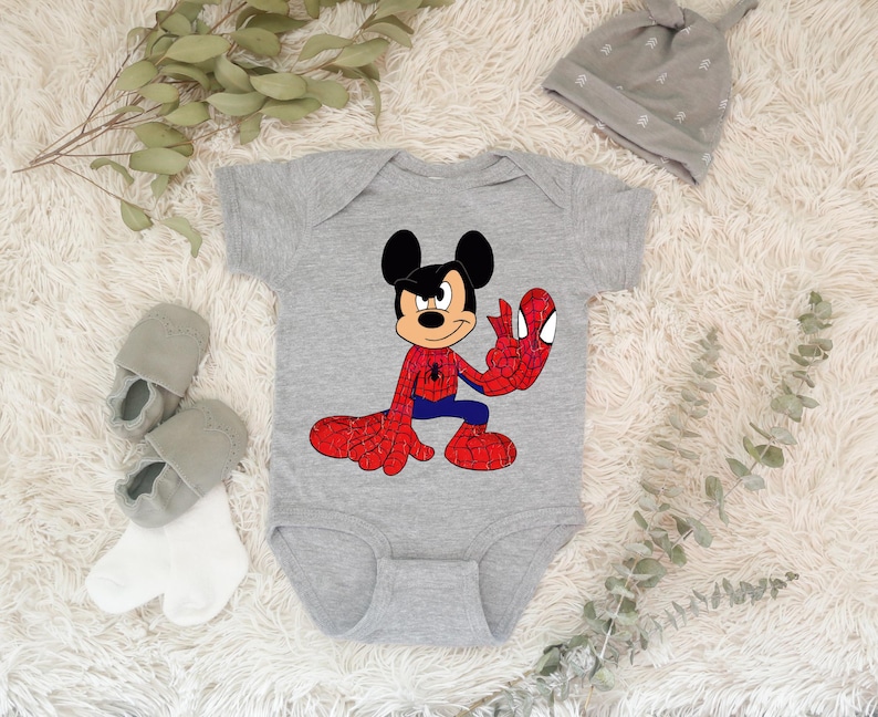 Op de afbeelding: Een grijze babybody met een cartoon Mickey Mouse-personage in een Spider-Man-kostuum. De body heeft korte mouwen en een drukknoopsluiting. Een grijze beanie, grijze babyschoentjes en witte sokken staan ook op de afbeelding.