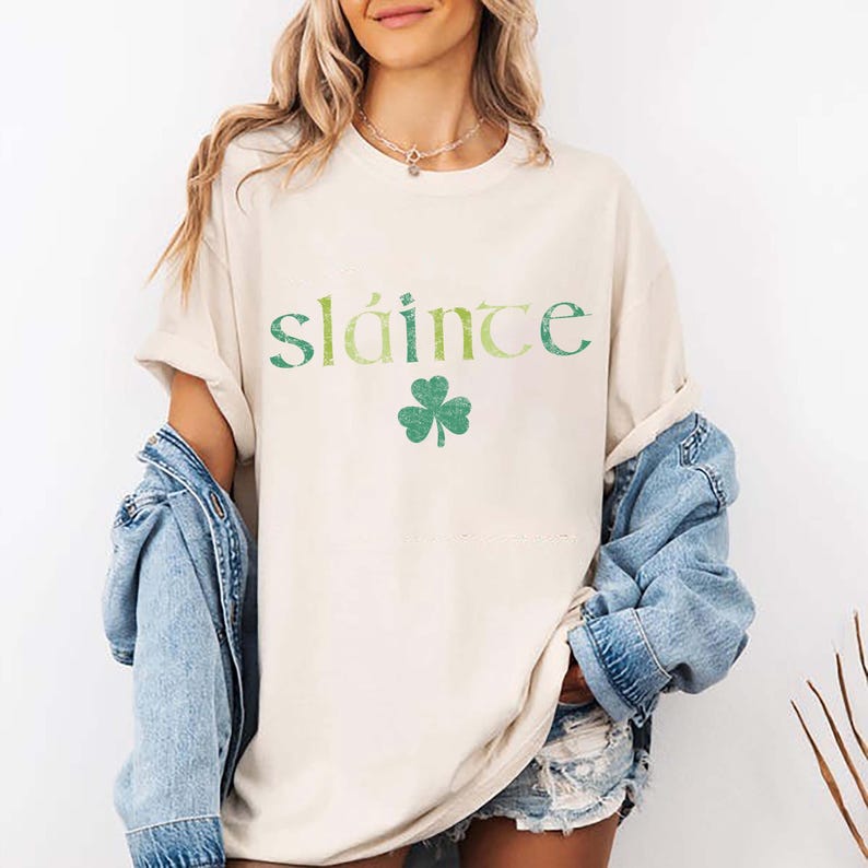May include: Cream-colored t-shirt with the word "sl&aacute;inte" in green lettering and a green shamrock. The shirt has rolled-up sleeves and is paired with a denim jacket. A necklace is visible.