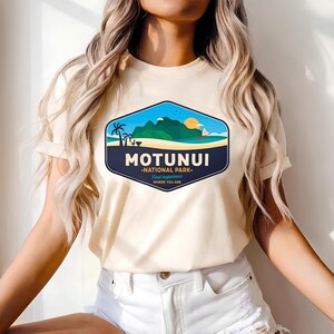 May include: A cream-coloured t-shirt displaying a graphic of a tropical landscape with the text "MOTUNUI NATIONAL PARK". The design includes a beach, mountains, palm trees, and a sailboat under a setting sun. The shirt also has the text "Find happiness where you are."