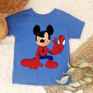 Op de afbeelding: Een blauw t-shirt met een stripfiguur die Mickey Mouse en Spider-Man combineert. Het personage draagt een Spider-Man-kostuum, met rode en blauwe kleuren. Het shirt ligt op een witte, zachte ondergrond.