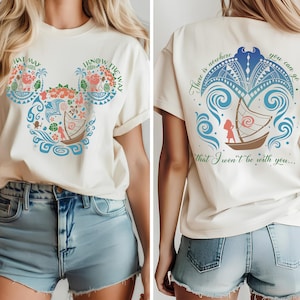 May include: Cream-colored t-shirt featuring a graphic design inspired by a Disney movie. The front displays a Mickey Mouse head shape filled with floral and nautical elements, with the text "HAT MAY" and "I KNOW THE WAY". The back has a boat design and the text "There is nowhere you can go that I won't be with you..."