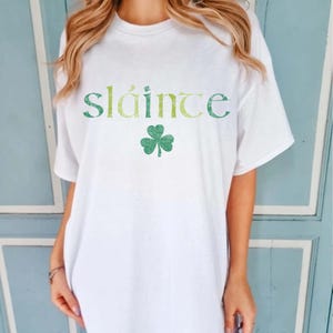 May include: White t-shirt with the word "Sl&aacute;inte" in green and yellow lettering and a green shamrock. The shirt is a casual style, suitable for St. Patrick's Day or Irish-themed events.