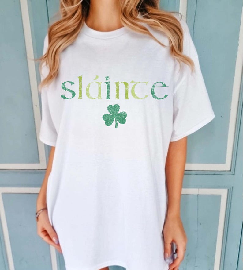 May include: White t-shirt with the word "Sl&aacute;inte" in green and yellow lettering and a green shamrock. The shirt is a casual style, suitable for St. Patrick's Day or Irish-themed events.