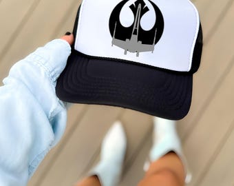May include: A black and white trucker hat featuring a black Rebel Alliance symbol graphic with a grey X-wing starfighter silhouette centered on the white foam front panel. The hat has a black brim and black mesh side and back panels.