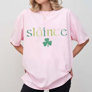 May include: A light pink t-shirt with the word "sl&aacute;inte" in green and yellow lettering and a green shamrock graphic. The shirt is a casual style.