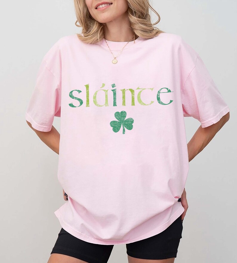 May include: A light pink t-shirt with the word "sl&aacute;inte" in green and yellow lettering and a green shamrock graphic. The shirt is a casual style.
