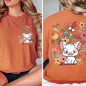 May include: Burnt orange t-shirt featuring a cartoon pig and rooster design with floral accents. The front of the shirt shows the pig and rooster with flowers. The back of the shirt has a larger version of the design surrounded by flowers.