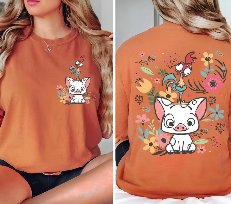 May include: Burnt orange t-shirt featuring a cartoon pig and rooster design with floral accents. The front of the shirt shows the pig and rooster with flowers. The back of the shirt has a larger version of the design surrounded by flowers.