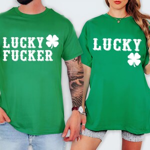 May include: Two green t-shirts with white text and a four-leaf clover design. One shirt says "LUCKY FUCKER" and the other says "LUCKY". The shirts are short-sleeved and worn by two people.