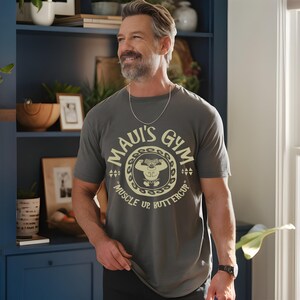 May include: A gray t-shirt with the text "Maui's Gym" in a circular design, featuring a cartoon weightlifter. The shirt also has the words "Muscle Up, Buttercup". The person is wearing a silver chain and a black watch.