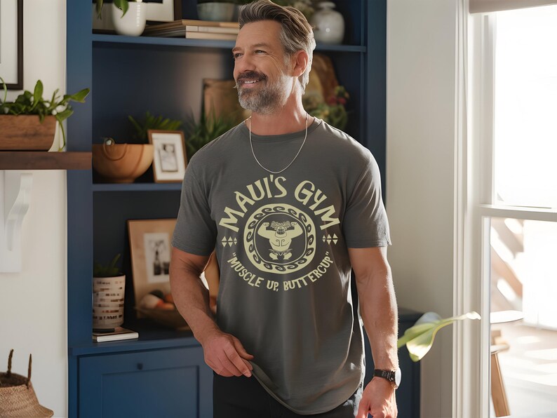 May include: A gray t-shirt with the text "Maui's Gym" in a circular design, featuring a cartoon weightlifter. The shirt also has the words "Muscle Up, Buttercup". The person is wearing a silver chain and a black watch.