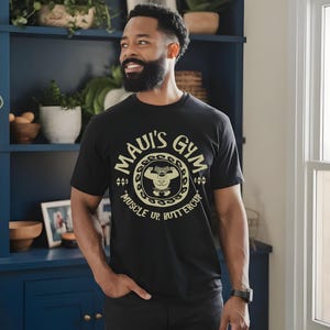 May include: Black t-shirt with the text "MAUI'S GYM" in a circular design, featuring a weightlifting cartoon character and the words "MUSCLE UP BUTTERCUP". The shirt is worn by a person.