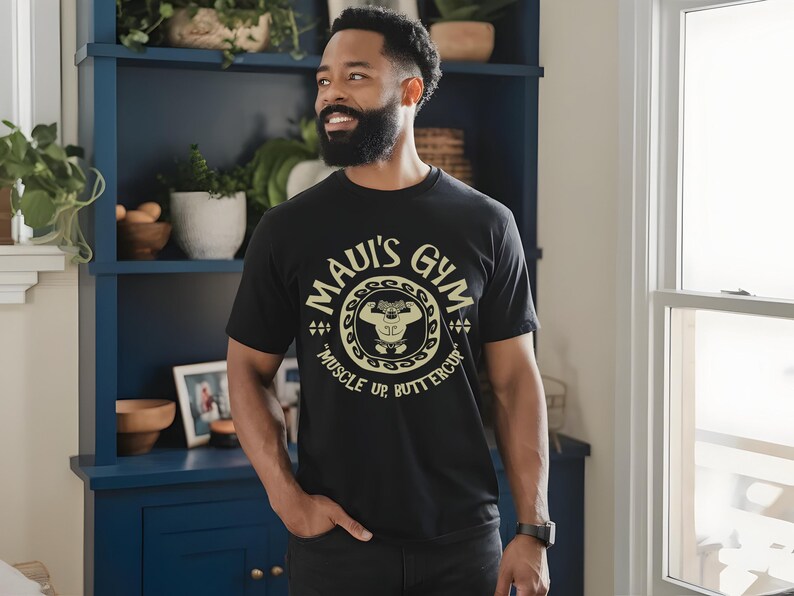 May include: Black t-shirt with the text "MAUI'S GYM" in a circular design, featuring a weightlifting cartoon character and the words "MUSCLE UP BUTTERCUP". The shirt is worn by a person.