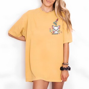 May include: Mustard yellow t-shirt featuring a cartoon graphic of a white pig-like character with a red and orange bird on its head, surrounded by flowers. The shirt has short sleeves and a relaxed fit.