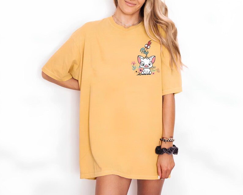 May include: Mustard yellow t-shirt featuring a cartoon graphic of a white pig-like character with a red and orange bird on its head, surrounded by flowers. The shirt has short sleeves and a relaxed fit.