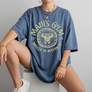 May include: A blue t-shirt with the text "MAUI'S GYM" in a circular design, featuring a cartoon weightlifter. The shirt also has the phrase "MUSCLE UP, BUTTERCUP" printed below the design. The shirt is a relaxed fit.