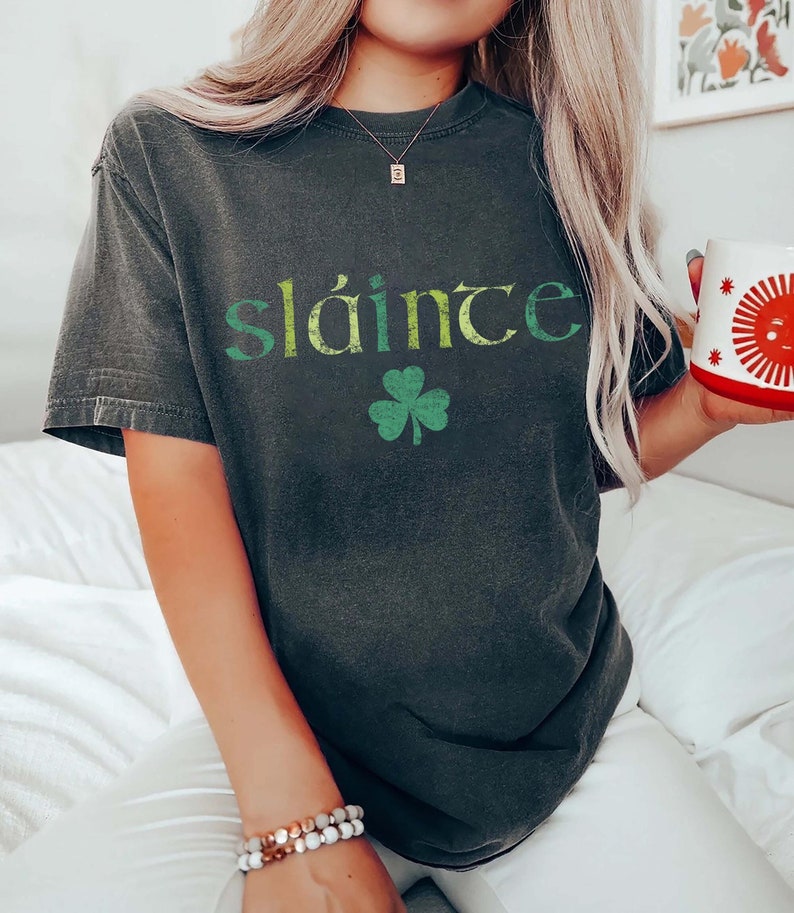 May include: Dark gray t-shirt with the word "Sl&aacute;inte" in green and yellow lettering, and a green shamrock. The shirt is a casual style.