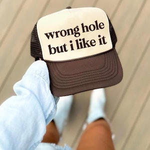 May include: A brown and white trucker hat with the text "wrong hole but I like it" in a bold, sans-serif font. The hat has a mesh back and a curved brim. The hat is being held up by a person.