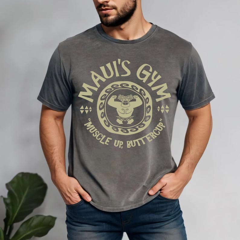 May include: A gray t-shirt with the text "MAUI'S GYM" in a circular design, featuring a muscular figure. The shirt also includes the phrase "MUSCLE UP, BUTTERCUP". The shirt is worn by a person with a beard and dark blue jeans.