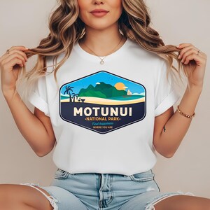 May include: White t-shirt with a graphic of a coastal scene, including the text "MOTUNUI NATIONAL PARK". The design features palm trees, a sailboat, and mountains under a blue sky and setting sun. The text "Find happiness where you are" is also included.