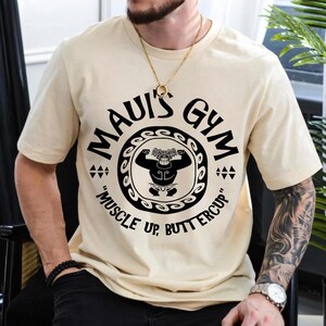 May include: Beige t-shirt with the text "MAUI'S GYM" in a circular design, featuring a muscular figure. The phrase "MUSCLE UP, BUTTERCUP" is below the design. The shirt has short sleeves and a crew neck.