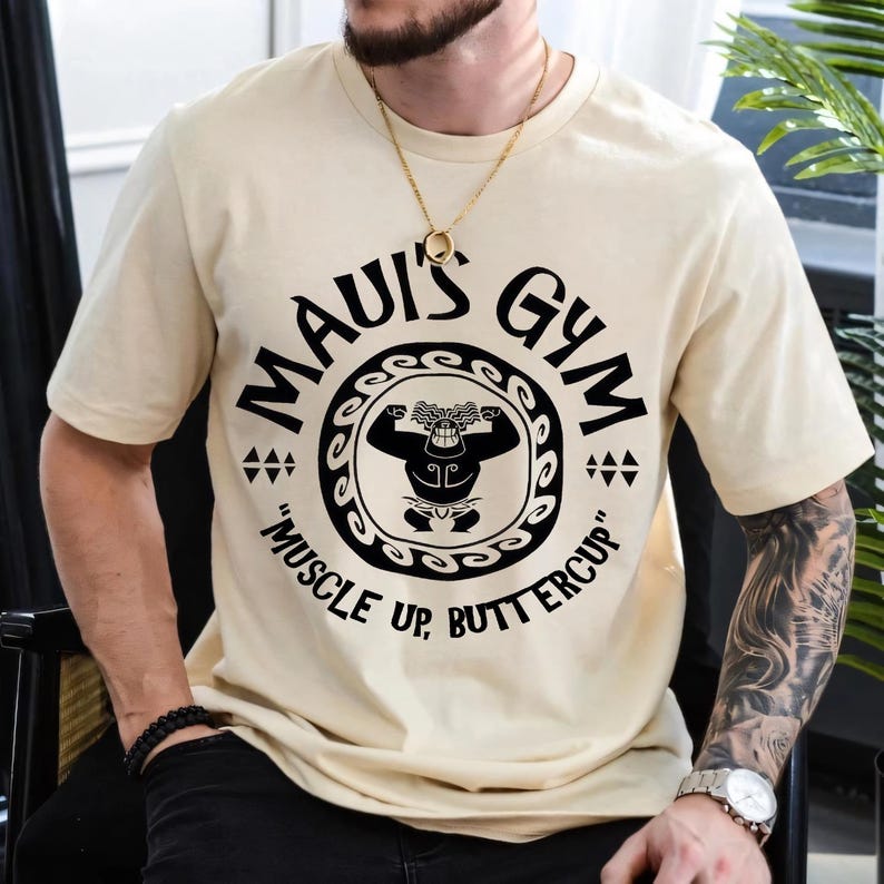 May include: Beige t-shirt with the text "MAUI'S GYM" in a circular design, featuring a muscular figure. The phrase "MUSCLE UP, BUTTERCUP" is below the design. The shirt has short sleeves and a crew neck.