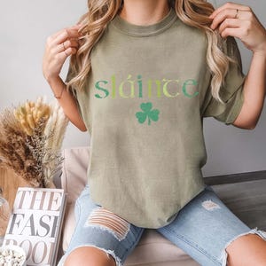 May include: Olive green t-shirt with the word "Sl&aacute;inte" in green and yellow lettering and a green shamrock graphic. The shirt is a casual style.