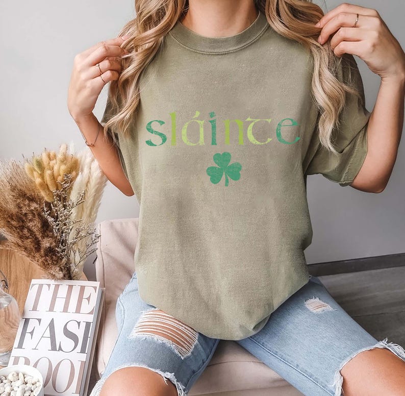 May include: Olive green t-shirt with the word "Sl&aacute;inte" in green and yellow lettering and a green shamrock graphic. The shirt is a casual style.