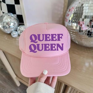 May include: Pink trucker hat with the text "QUEEF QUEEN" in purple. The hat has a mesh back and a curved brim. The hat is held up by a hand. The background includes disco balls.