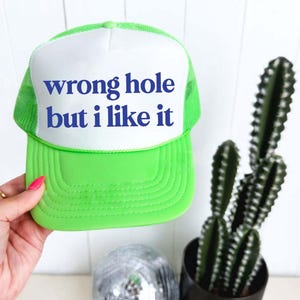 May include: A neon green trucker hat with a white front panel. The text "wrong hole but i like it" is printed in blue on the front. The hat is held by a person's hand. A cactus and disco ball are in the background.